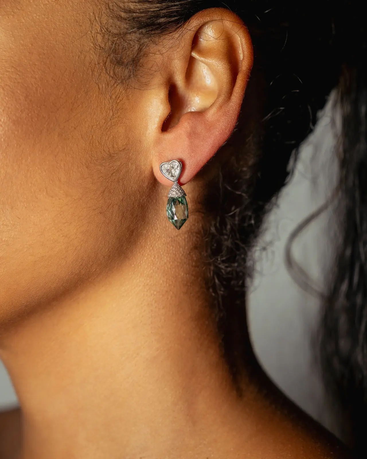 A heart-shaped diamond earring with a faceted green drop, worn